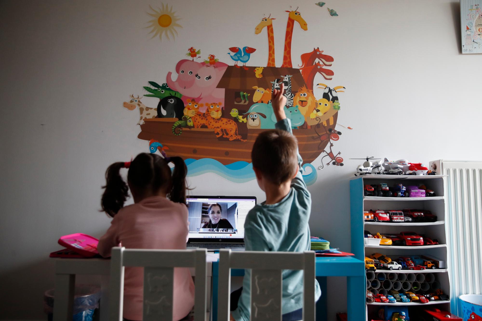 A kindergarten teacher reads a fairy story as Panos raising his hand with his twin sister Marina watch the online lesson in Athens, Thursday, Nov. 19, 2020. Most other European countries have vowed to keep schools open, but the pandemic has hit Greece hard for the first time in recent weeks following a successful lockdown in the spring, overwhelming hospitals in parts of the country. State television is making and broadcasting lessons, while teachers sit in empty classrooms talking to remote students. Despite some problems, they say it keeps children in touch with their schools. (AP Photo/Thanassis Stavrakis)