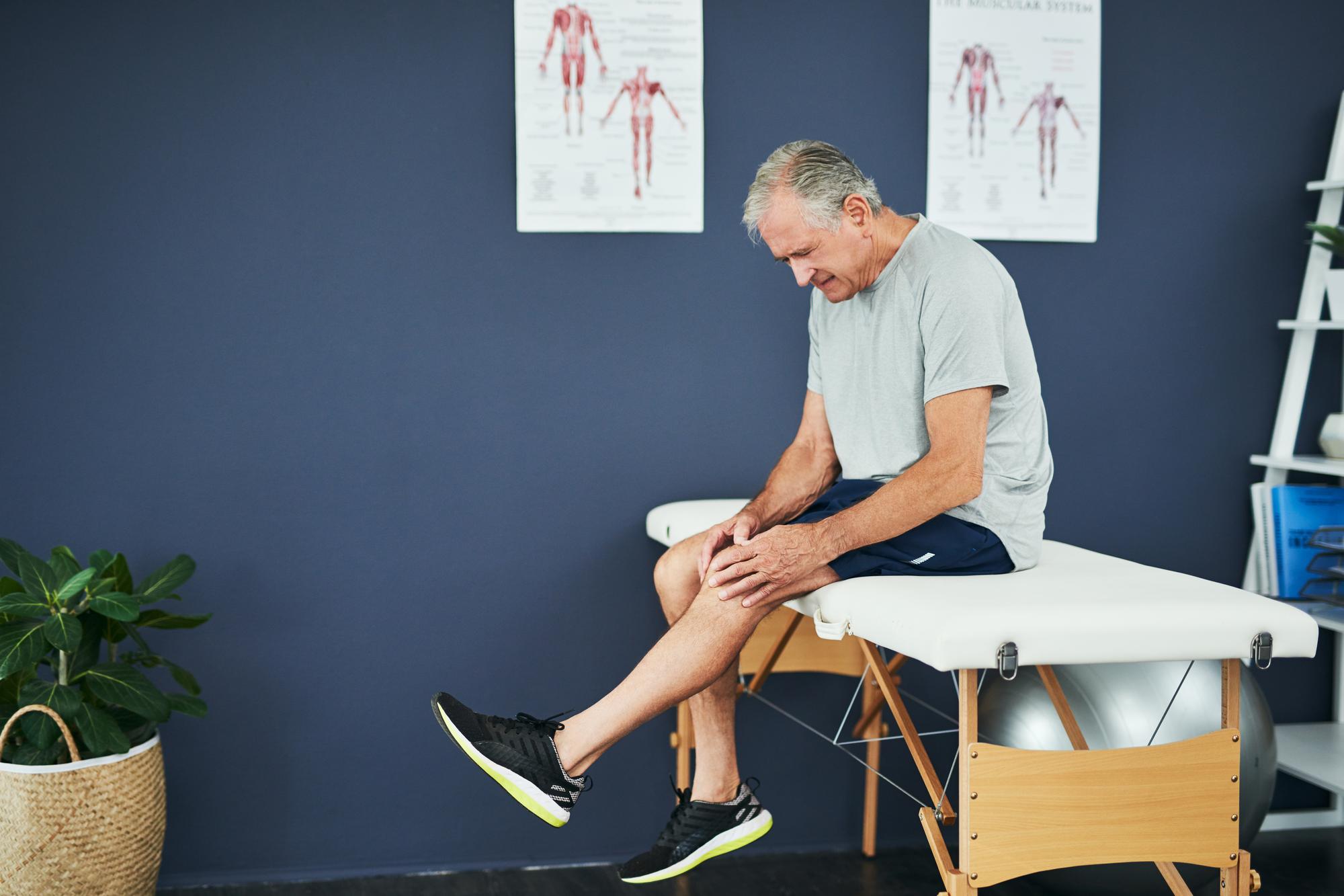 Cropped shot of an handsome senior man holding his knee while sitting on the bed in a physiotherapy office