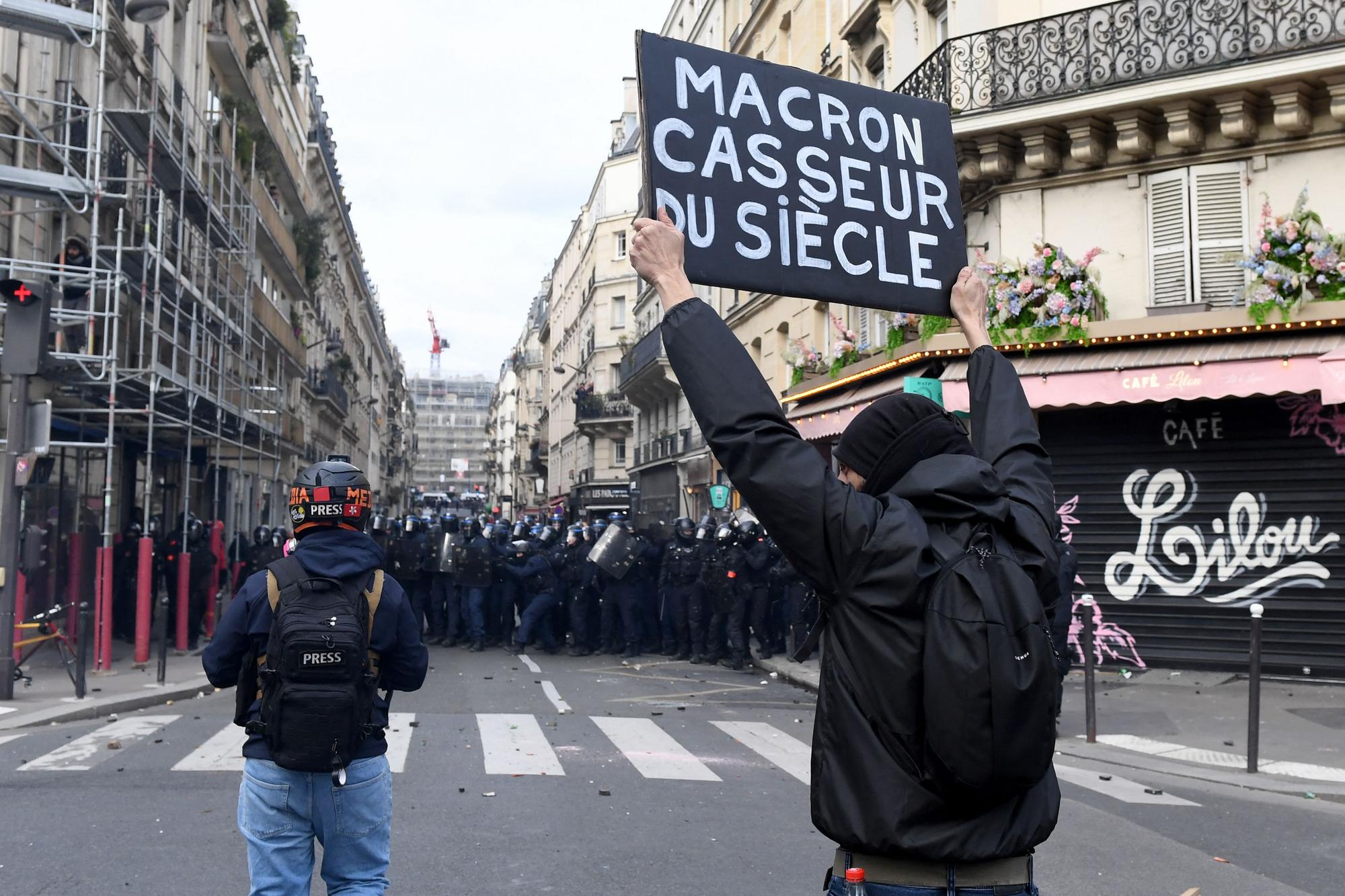 A man holds a placard reading in French "Macron, thug of the century" in front of police forces during a demonstration in Paris on March 7, 2023, as part of a nationwide day of strikes and protests called by unions over the proposed pensions overhaul, which include raising the minimum retirement age to 64 from 62 and increasing the number of years people have to make contributions for a full pension. (Photo by Alain JOCARD / AFP)