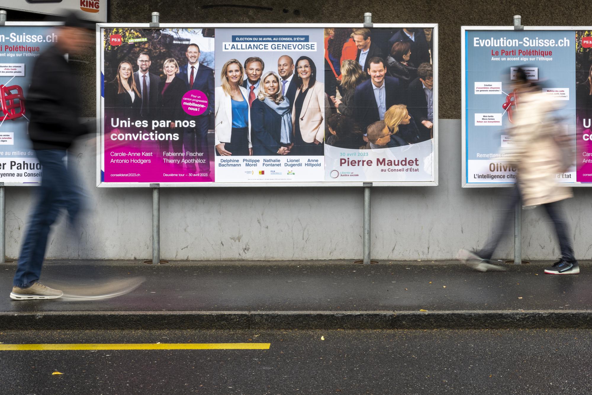 Deux personnes passent devant les affiches electorales du deuxieme tour de l'election du Conseil d'Etat, l'affiche du parti Socialiste et les Verts avec les candidats, Carole-Anne Kast, Antonio Hodgers, Fabienne Fischer, Thierry Apotheloz, l'affiche de l'alliance genevoise avec les candidats, Delphine Bachmann - Le Centre, Philippe Morel - MCG, Nathalie Fontanet - PLR, Lionel Dugerdil - UDC et Anne Hiltpold - PLR et l'affiche de Libertes et Justice sociale Pierre Maudet, ce jeudi 20 avril 2023 a Geneve. Les citoyens genevois voteront pour l'election du second tour de l'election du Conseil d'Etat du dimanche 30 avril 2023. (KEYSTONE/Martial Trezzini)
