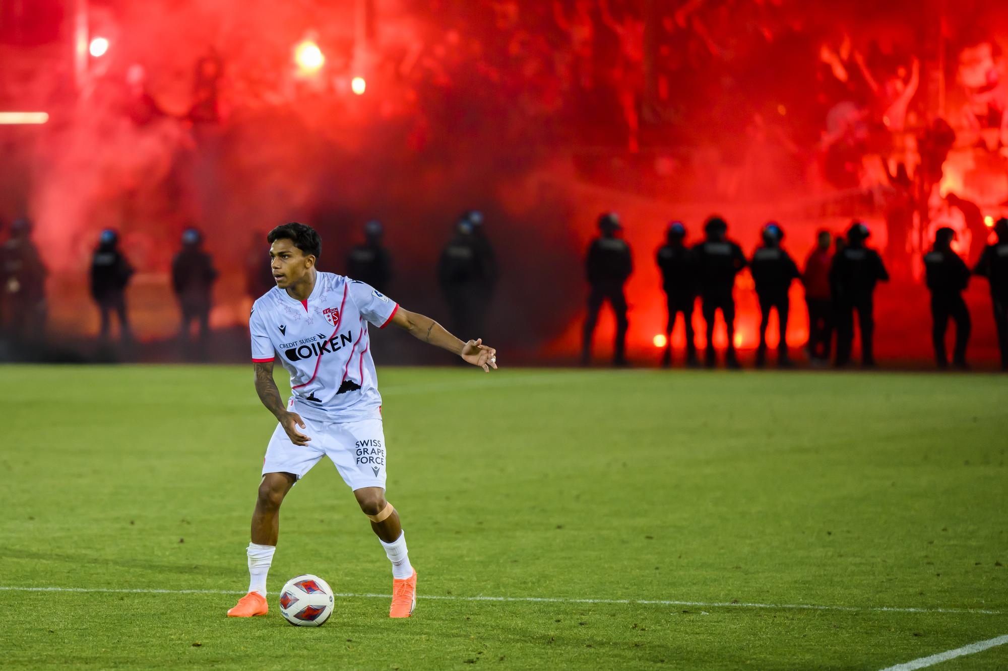 Les supporters valaisans allument des fumigenes davant les policiers anti-emeute de la police vaudoise et le milieu valaisan Baltazar Costa lors du match retour du barrage de promotion en Super League et de relegation en Challenge League de football entre le FC Stade Lausanne Ouchy, SLO, et le FC Sion ce mardi 6 juin 2023 au stade olympique de la Pontaise a Lausanne. (KEYSTONE/Jean-Christophe Bott)