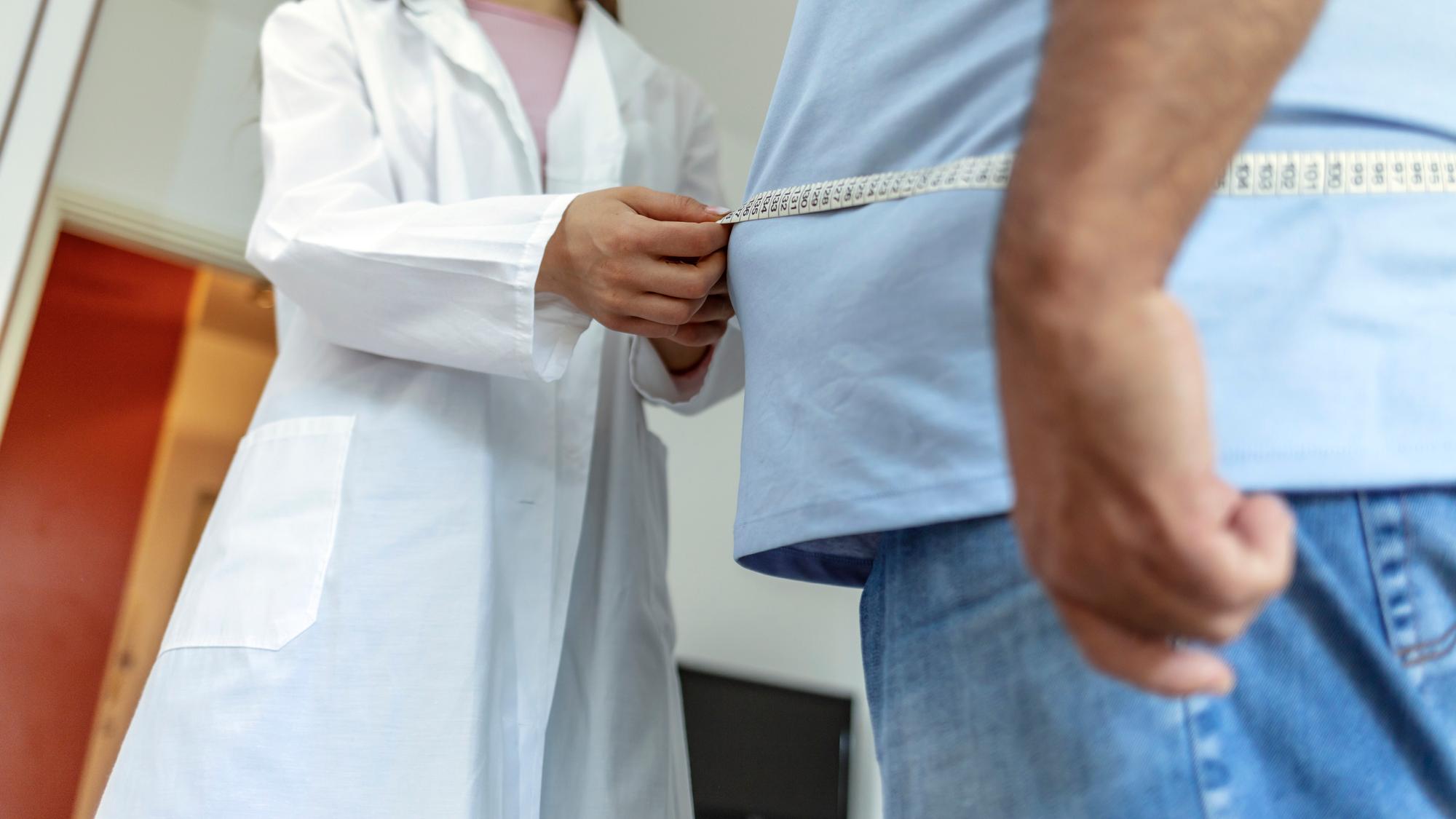 A nutritionist doctor measures the body of a male patient with a measuring tape on adipose tissue and excess weight. Overweight obesity man seeing doctor for treatment of his illness and weight loss.