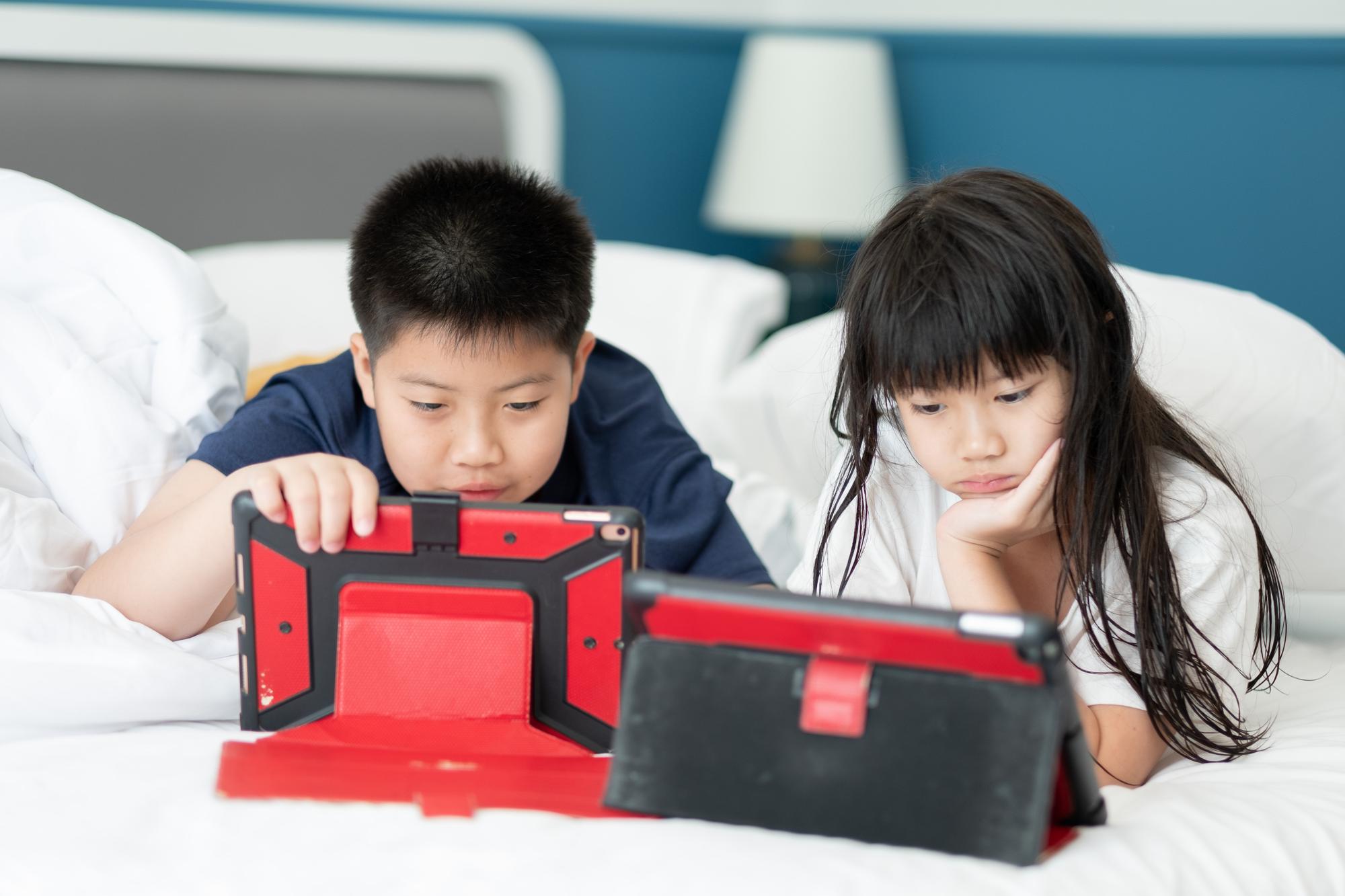two chinese children addicted tablet, asian child watching telephone together on their bed, kid using smartphone