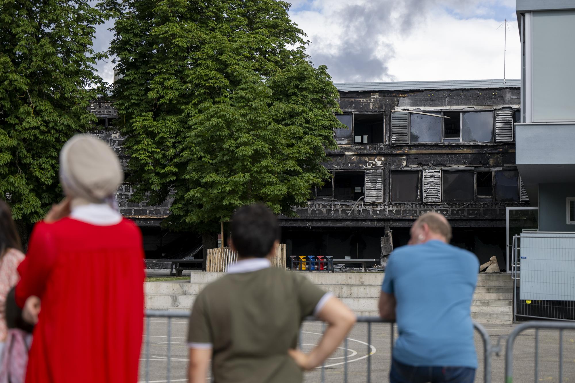 Des personnes observent la facade de l'ecole des Ranches detruite par les flammes, ce mercredi 5 juillet 2023 a Vernier pres de Geneve. Des degats importants apres l'incendie qui s'est declare dans la nuit de mardi a mercredi, une 40 de pompiers du Service d'incendie et de secours (SIS) et de pompiers volontaires etait sur place afin de maitriser le sinistre. (KEYSTONE/Martial Trezzini)