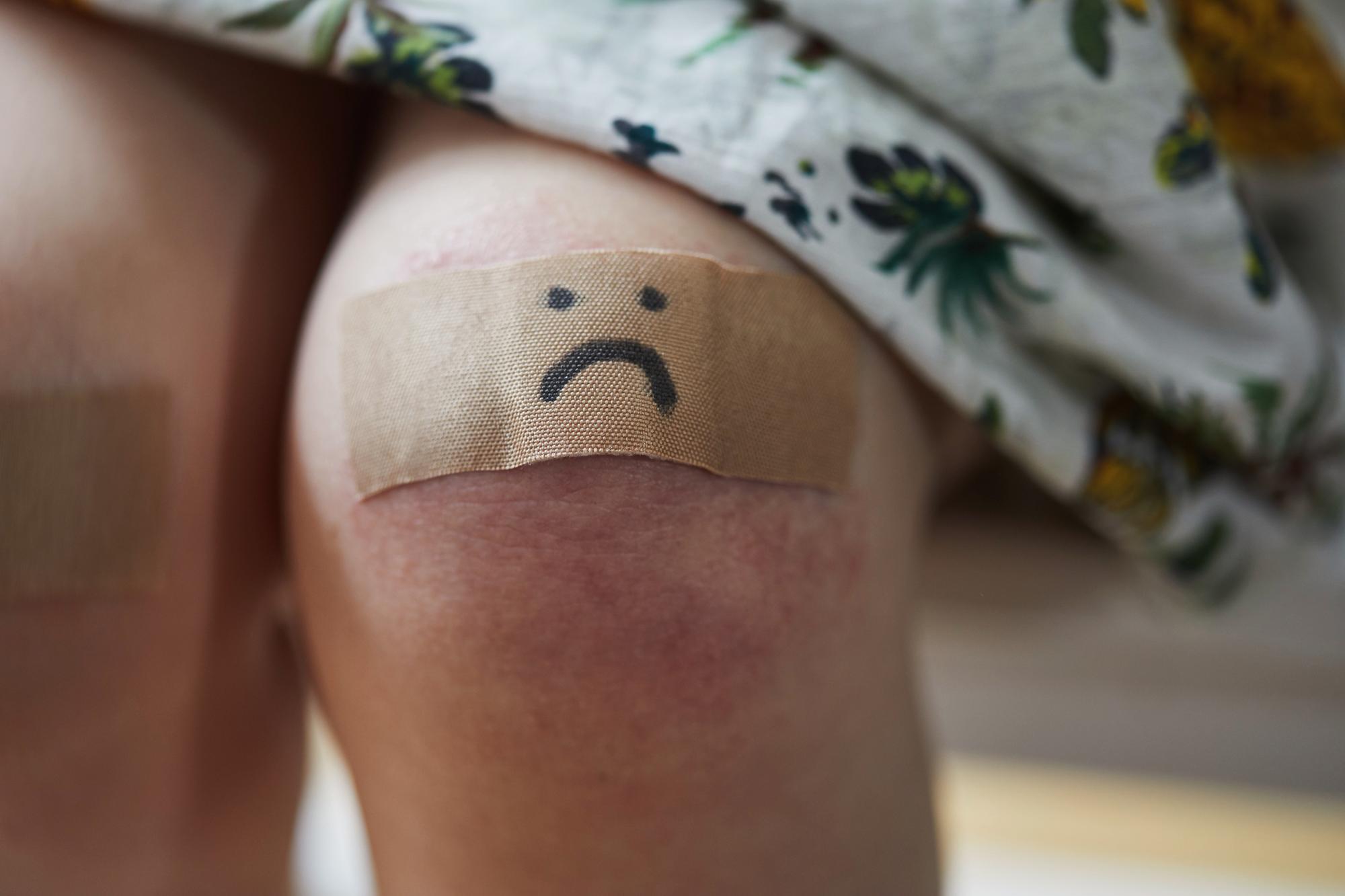 Toddler girl with a plaster on her grazed knee with a sad face drawn on it