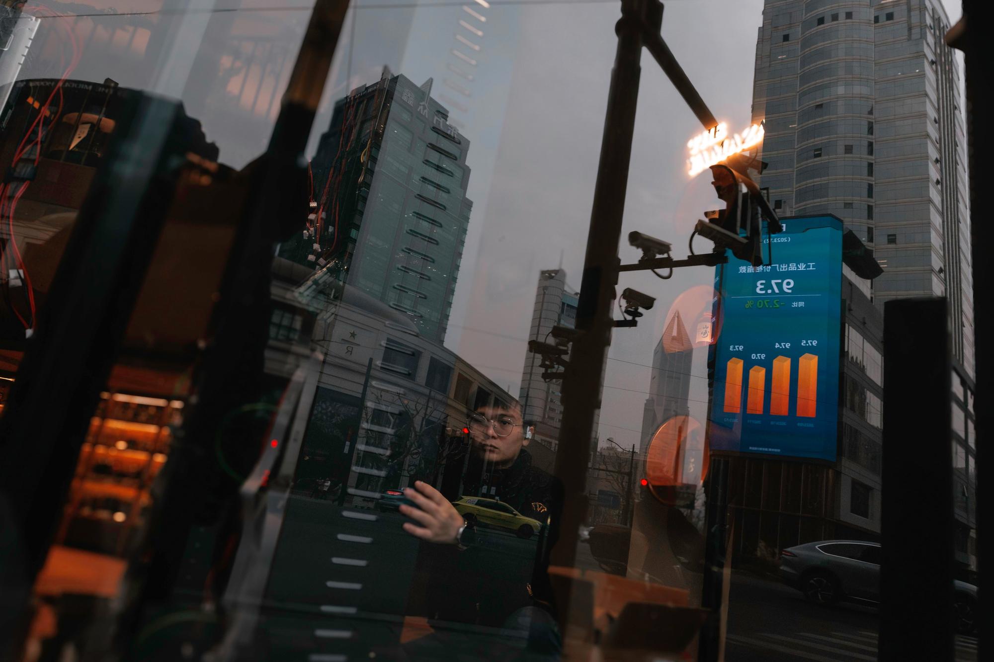 epa11112042 A man works in a shop while a large screen showing the latest stock exchange and economic data is seen reflected on the shop's window in Shanghai, China, 29 January 2024. A Hong Kong court has ordered China Evergrande to liquidate after a hearing with the real-estate company who has around 300 billion dollars worth of debt. EPA/ALEX PLAVEVSKI